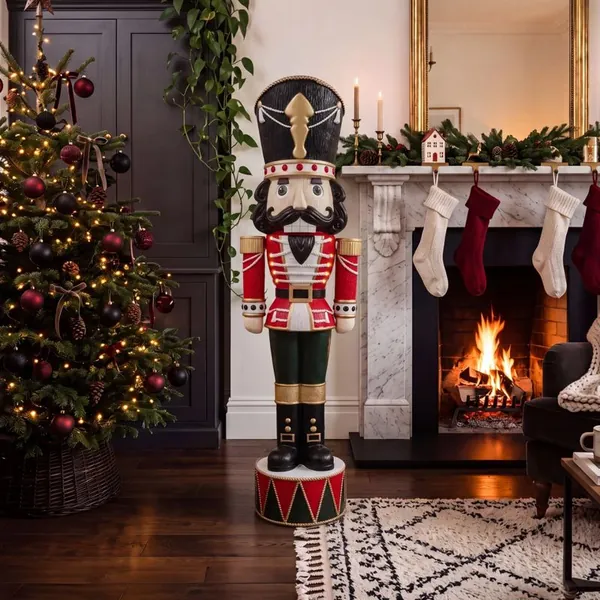 Traditional Festive Otto nutcracker in red and green beside a Christmas tree and fireplace